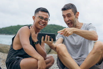 Couple of male friends having big laughs sitting on rocks by the sea looking at cellphone. Filipino young man and caucasian guy enjoying social media on smart phone. Ethnicity diversity, sharing jokes