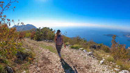 Woman with backpack on idyllic hiking trail along the Budvanian Riviera from Sveti Stefan to Goli Vrh, Montenegro, Balkan, Europe. Looking at touristic coastal town Budva and Sveti Nikola island