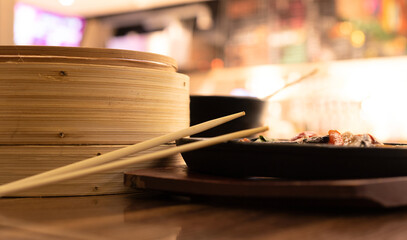 Fototapeta na wymiar Chopsticks, delicious asian food in cast-iron pan and bamboo steamer on wooden background. Selective focus. Asian food concept