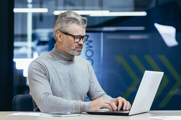 Serious thinking businessman inside office at work with laptop, senior gray haired man working sitting at desk.