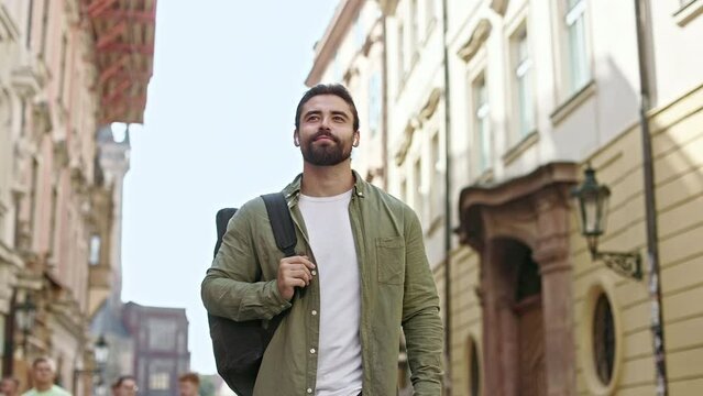 Side view of sandsome, brunette man with beard traveling, walking down street. Male tourist with earphones, carrying backpack, admiring buildings, enjoying. Concept of traveling.