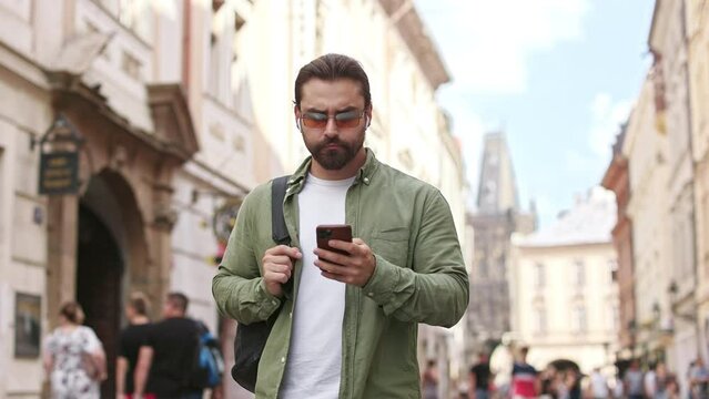 Side view of busy tourist carrying backpack, gong walking down street. Handsome male with beard wearing sunglasses, holding, using smartphone. Concept of modern lifestyle.