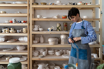 Creative artisan crafts woman tying apron and preparing for pottery master class in her own art studio. Relaxed lady in good mood started her work day. Small art business concept