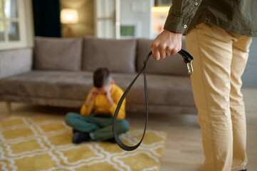 Closeup of father holding leather belt to punish crying son sitting on floor