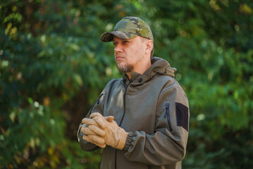 Fototapeta premium Mature man dressed in military uniform, camouflage cap and tactical gloves clasp his fingers both hands over the forest green background. a man dressed in green uniform. Tactical dress Concept.