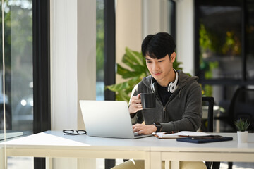Handsome asian male freelancer drinking coffee and working online from home on laptop computer