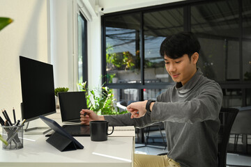 Smiling asian male freelancer looking at wristwatch to managing time during working online in modern home office