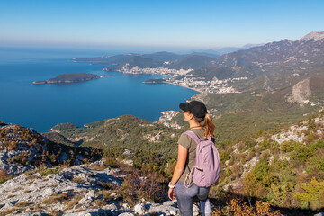 Hiking woman with backpack enjoying panoramic aerial view on coastline of Budva and Sveti Nikola Island seen from Goli Vrh, Adriatic Mediterranean Sea, Montenegro, Balkan, Europe. Budvanian Riviera