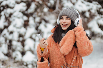 Pretty young long-haired woman in pink coat enjoy winter day and listening music with headphones at snowy park
