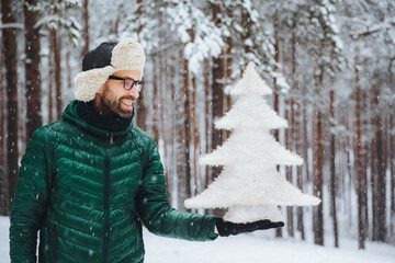 Smiling middle aged male looks with happy expression at artificial white small fir tree, going to celebrate New Year`s holidays, spends morning on fresh air in forest during snowy weather