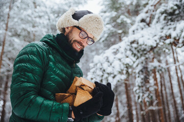 Pleased man in spectacles looks hapily at firewood which he log in winter forest, going to make fire, wears green anorak, poses against white winter forest. People, lifestyle, recreation concept