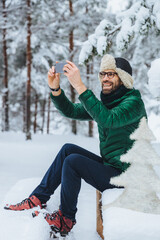 Vertical portrait of pleasant looking male makes photo with smart phone, makes shots of beautiful winter landscapes, enjoys nature and frozy weather, has pleasant smile. People, technology concept