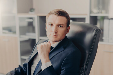 Attractive male manager sits in chair at his cabinet, dressed formally, rests for minute after hard...