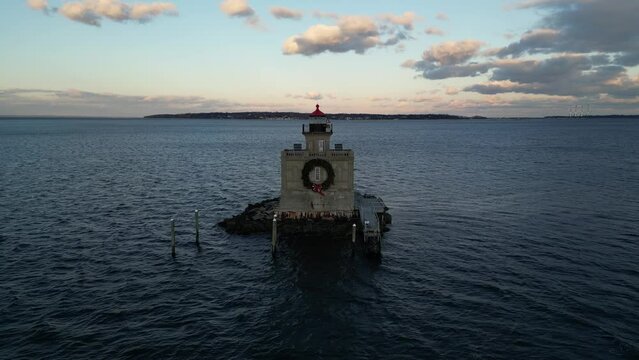 An aerial view of the Huntington Harbor Lighthouse on Long Island, NY at sunset, with a Christmas wreath. The stationary camera is in front of the lighthouse with beautiful clouds and a blue sky.