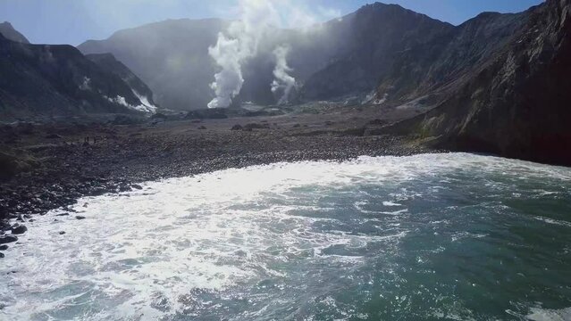 Flying towards Whakaari White Island crater volcano, small volcanic pebble beach, aerial