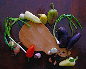 onion, garlic, sweet pepper, eggplant scattered on a dark wooden surface, around a cutting board,...