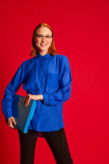 Portrait of young redhead girl in blue shirt and glasses posing with laptop over red background. Happy worker