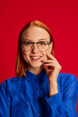 Portrait of young redhead girl in blue shirt and glasses posing over red background. Motivated employee