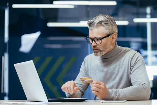 Senior Gray Haired Businessman In Office Frustrated And Sad Trying To Make Bank Transaction And Purchase In Online Internet Store, Man Holding Bank Credit Card And Using Laptop.