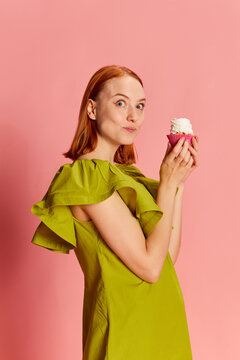Portrait of young beautiful girl in cute dress posing with cupcake over pink background. Sweets lover