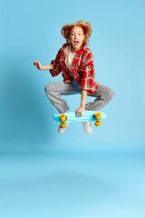 Portrait of young redhead girl in casual checkered shirt posing, jumping with skateboard over blue background