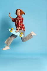 Portrait of young redhead girl in casual checkered shirt posing, running with skateboard over blue background