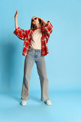 Portrait of young redhead girl in casual checkered shirt listening to music in headphones and dancing over blue background