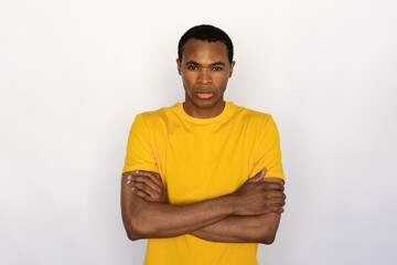Fototapeta premium Portrait of serious young man posing with folded arms against white background. African American guy wearing yellow T-shirt looking at camera. Confidence concept