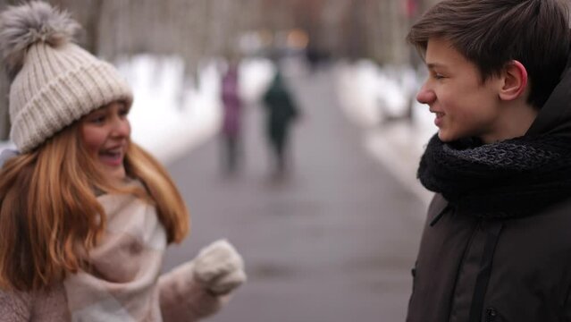 Thoughtful teen boy standing on park alley as joyful girl covering eyes with hands surprising friend. Portrait of handsome boy and beautiful girl meeting on winter day outdoors leaving talking smiling