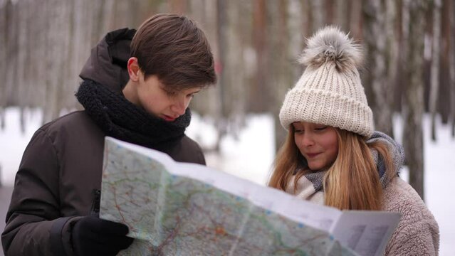 Teenage boy and girl talking examining paper map standing in winter park. Portrait of concentrated Caucasian teen couple planning vacations dating outdoors. Lifestyle and leisure concept
