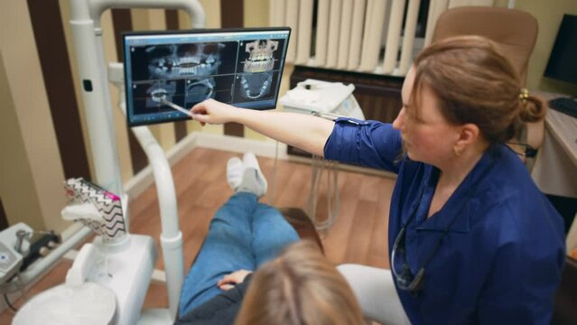 Dentist talking, gesturing and showing results of x-ray teeth on monitor screen to patient in dental chair. Cabinet in tooth clinic. Preventive examination. Treatment, healthcare and medicine.