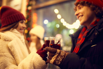 Young couple in love walking at festive Christmas street and drinking mulled wine. Concept of...