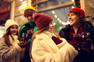 Happy young men and girls, friends walking at festive Christmas street and drinking mulled wine....