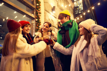 Fototapeta premium Happy young men and girls, friends walking at festive Christmas street and drinking mulled wine. Concept of holidays, winter, New Years fairs, friendship
