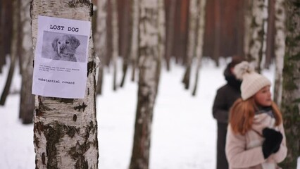 Missing pet placard on tree with blurred teen girl and boy at background looking for dog in winter forest. Anxious worried Caucasian friends searching in park outdoors