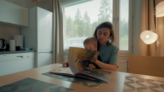Caring mother shows little son bright pictures in book sitting at kitchen table. Woman and toddler boy spend weekend morning in big cottage house