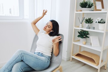 Woman sitting in a chair listening to an audiobook on wireless headphones at home in jeans and a white T-shirt, fall lifestyle comfort