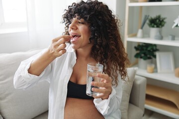 Pregnant woman smile blogger takes vitamins and medicines pills with a glass of water close-up sitting on the sofa at home freelancer in the last month of pregnancy lifestyle before childbirth