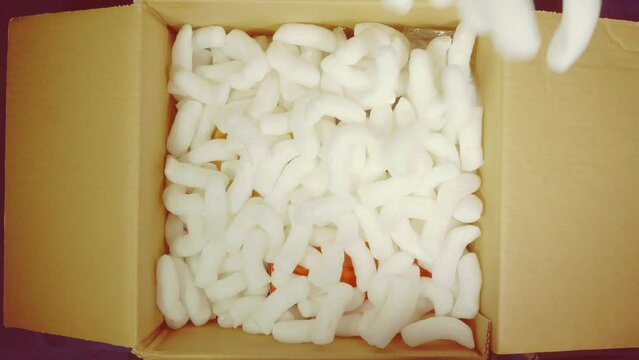 Closeup overhead shot of items in a cardboard box, with loosefill polystyrene protective packing, being poured over the top, ready for shipping / posting.