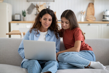 Modern family mother and daughter watching movie online looking at laptop screen while resting together on sofa at home. Mom and teen girl child shopping via internet, sitting on couch using computer