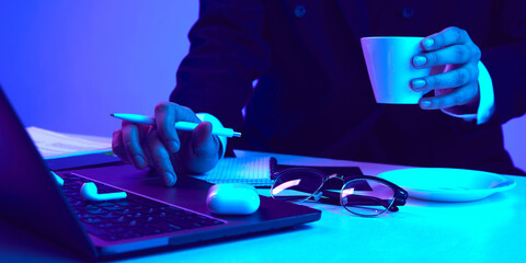 Making notes on laptop and drinking coffee. Working desk with computer, notebook, headphones and phone in neon colored light