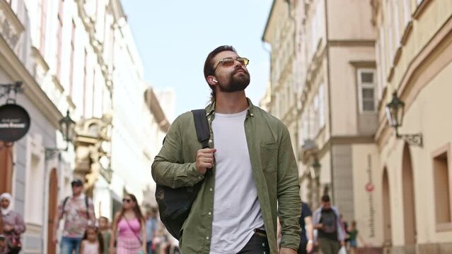 Front view of male tourist traveling, admiring, watching architecture, buildings, smiling. Handsome man with beard carrying backpack, listening to music. Concept of tourism.