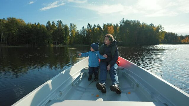 Mother and little son sit in boat drifting on lake near lush autumn forest. Woman shows toddler beautiful nature of countryside on sunny day