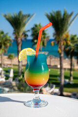 Colourful cold Rainbow Paradise cocktail drink served in glass at pool bar overlooking blue pool, sea and palm trees, relax and holidays at sea