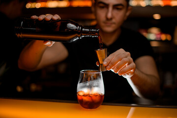 Close-up of bottle and jigger in hands of male bartender and glass with iced drink.