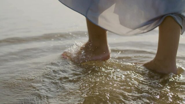 Legs closeup of woman with red nails walking in sea water. Young barefoot Caucasian female strolling on the beach, stepping on wet sand while water hardly reaching her ankles. Nature, holiday concept.