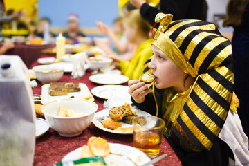 a boy dressed as a pharaoh at a children's party sits at a table with cookies, muffins and cakes and eats a pie. © Ruslan Russland