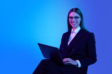 Woman in stylish official suit working on laptop over blue background in neon light. Successful...