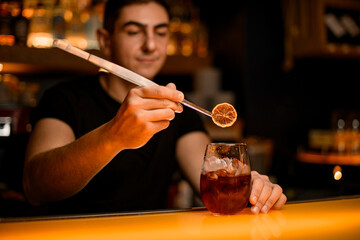 view on male hand holding tweezers with orange slice over glass with cold cocktail