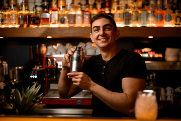 Authentic smiling male barman in bar interior with shaker making alcohol beverages